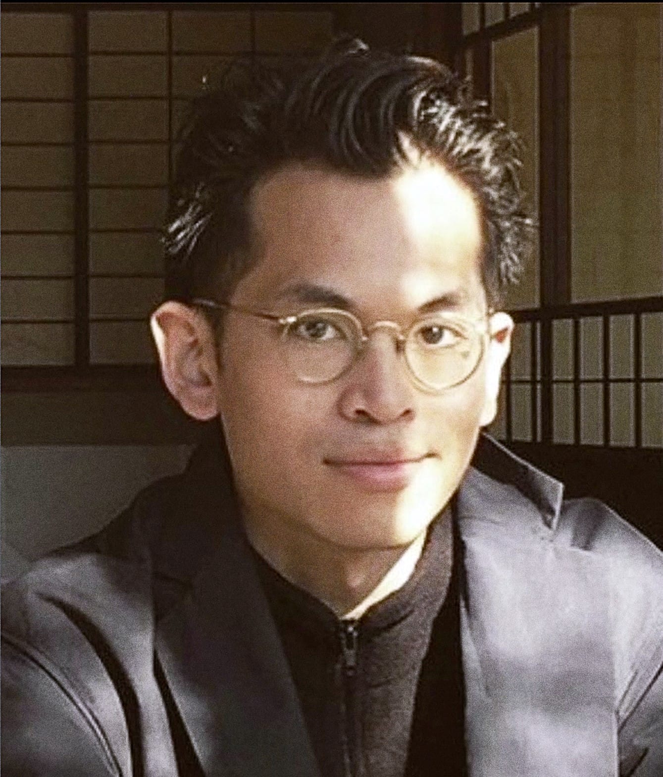 portrait of a young man with short dark hair, circular glasses, and a coat in front of a Japanese screen smiling