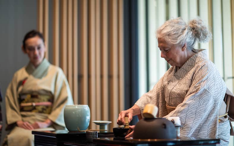 Two women sit in kimonos on tatami while doing a presentation of Japanese tea ceremony.
