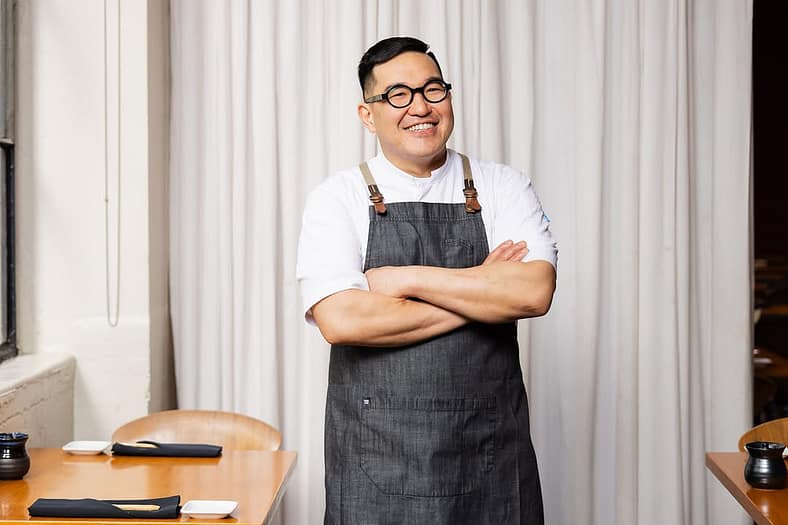 Chef Masu sushi standing with arms crossed wearing chef's apron in his Masu Sushi restaurant.