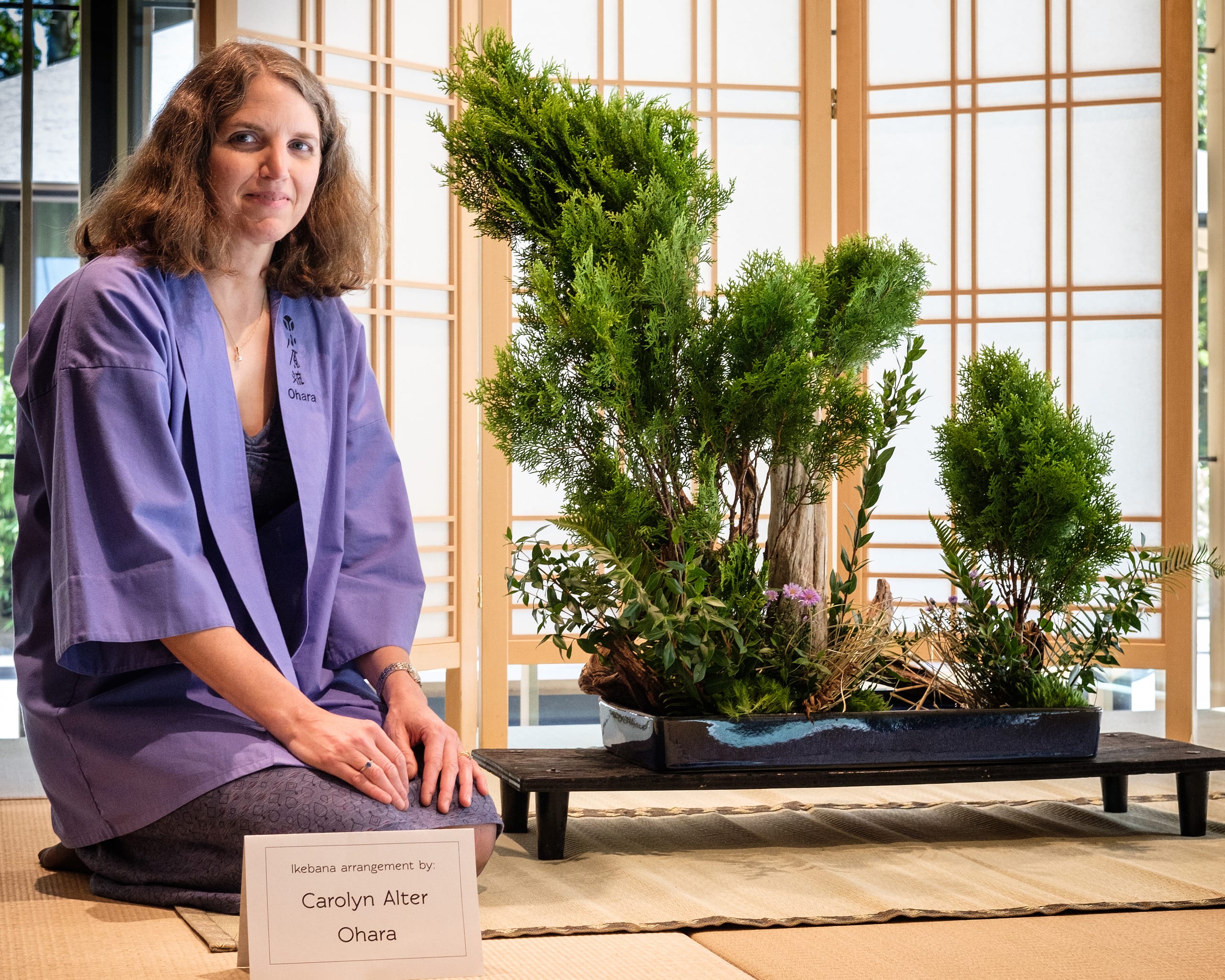 A woman sits next to ikebana.