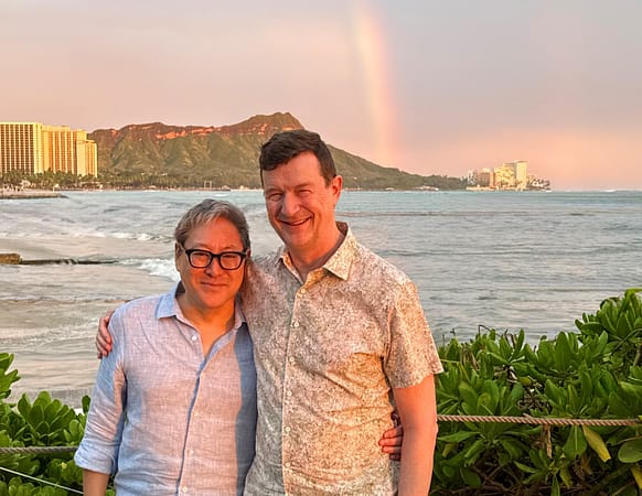 a couple stands in front of a body of water with a rainbow dipping down from the sky