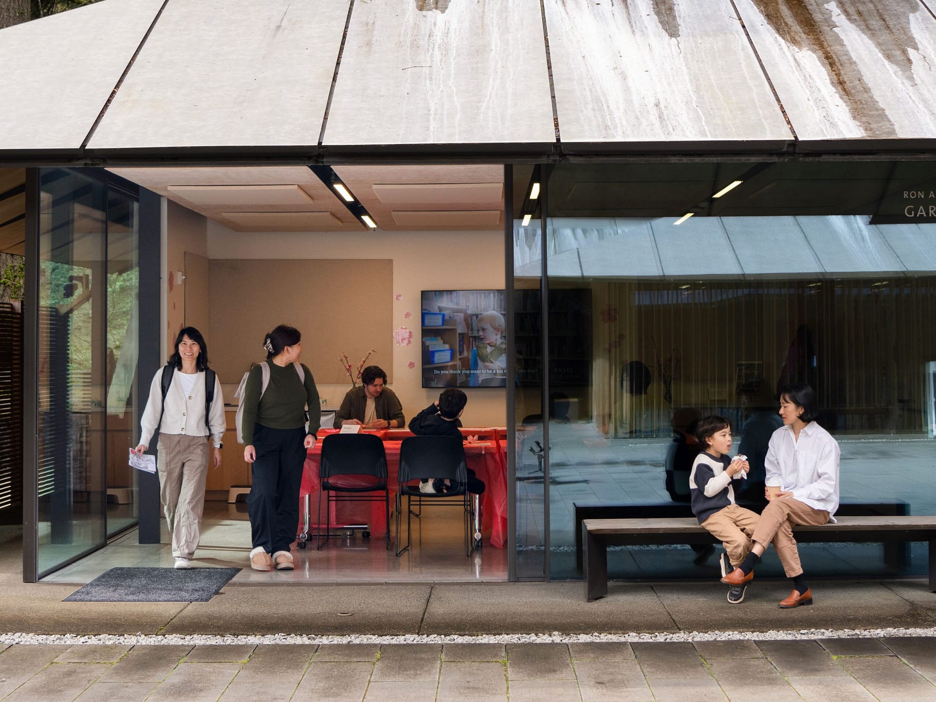 Mother and son sitting outside of glass building with other persons sitting inside and exiting the building. Instructional video on TV inside of building.