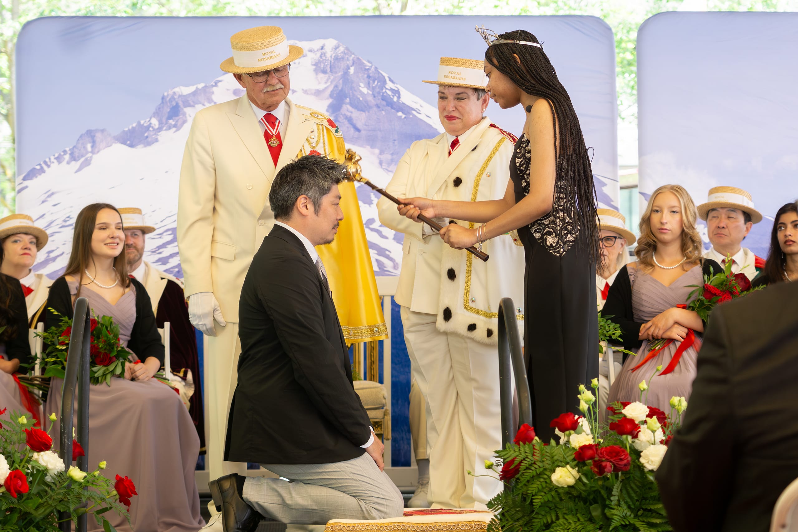 A man kneels while being knighted by a young woman wearing a crown and holding a scepter.