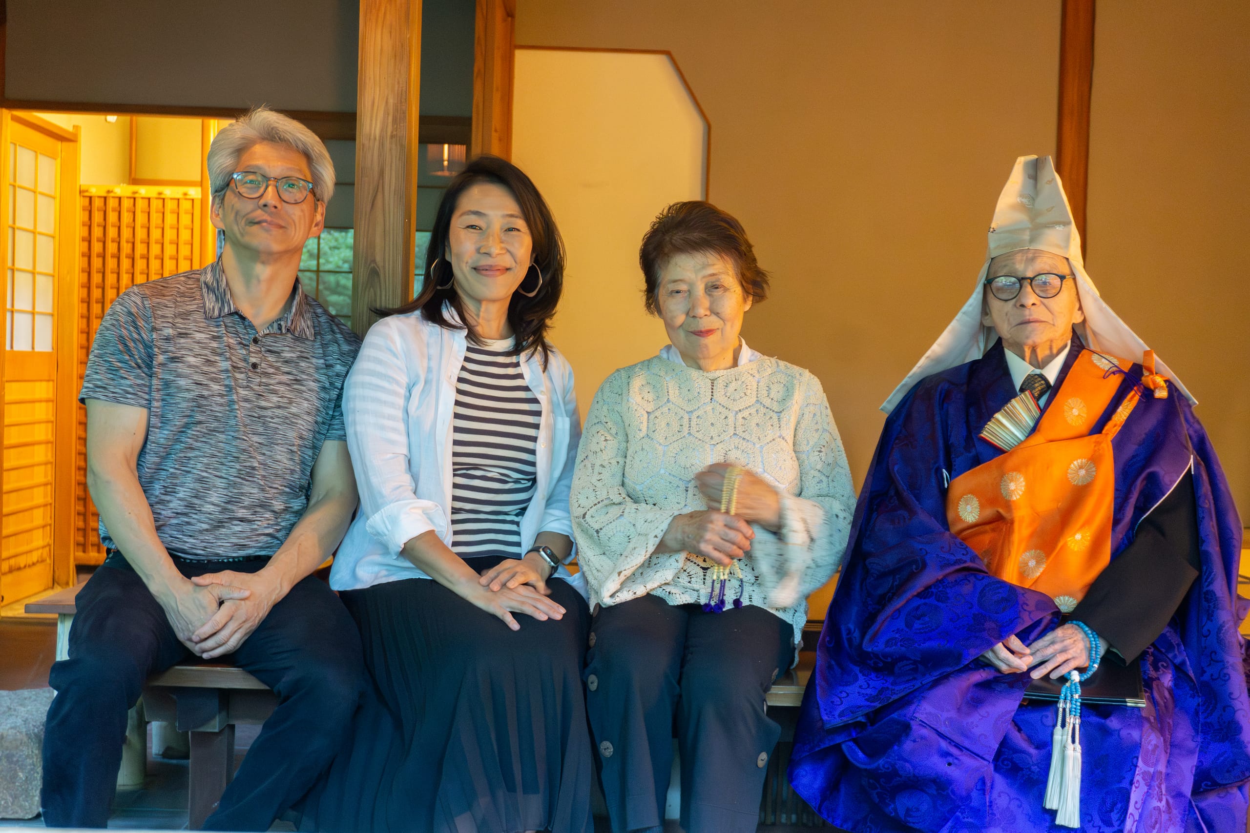 four Japanese people sitting in a tea house: a man in a grey shirt with white hair, woman in striped shirt with black hair, older woman in white shirt and auburn hair holding prayer beads, and an older man with a Buddhist reverend outfit (white hat, purple kimono, and orange cover) holding prayer beads