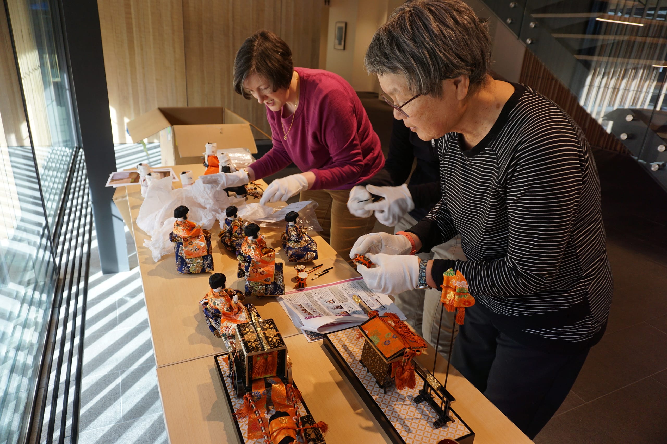 a female garden volunteer and a female staff member wear white gloves to set out Japanese dolls on a long table