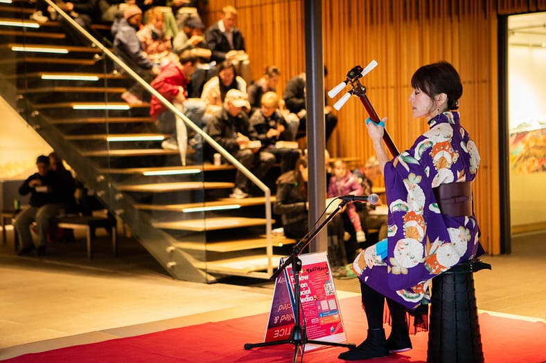 a woman sits on a stool while wearing a kimono playing a three stringed shamisen in front of a microphone and seated crowd