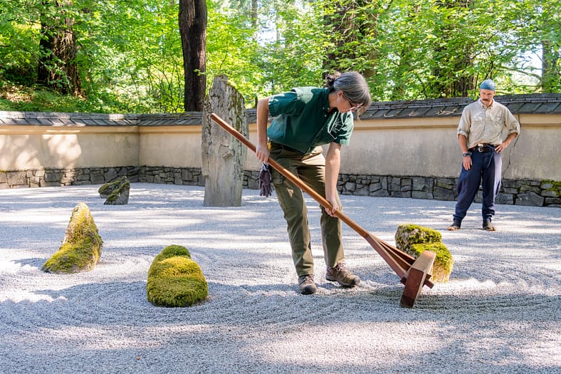 a woman rakes gravel in a Japanese garden while another gardener looks on approvingly