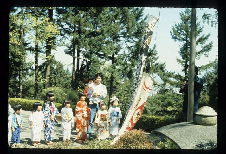 Children look at a cloth carp streamer before it's hoisted into the air on a flag pole.