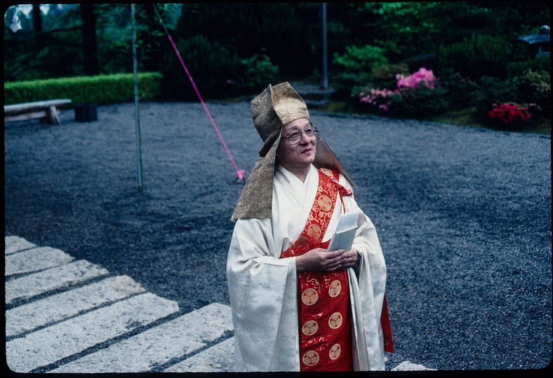 a man in a white kimono stands in a gravel courtyard with a prayer book