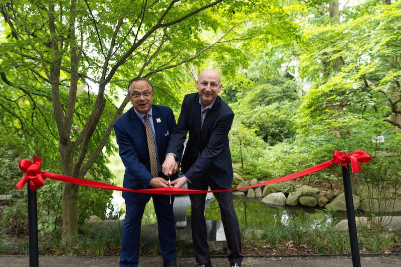two men stand with scissors poised to cut a red ribbon in a lush garden