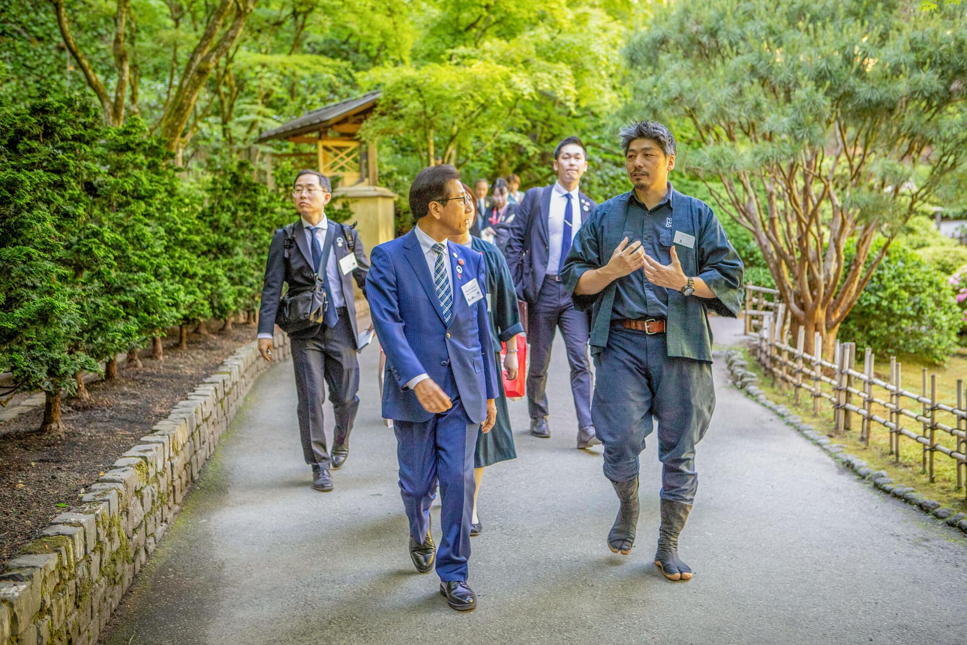 two men talk and walk in a garden leading a group of people