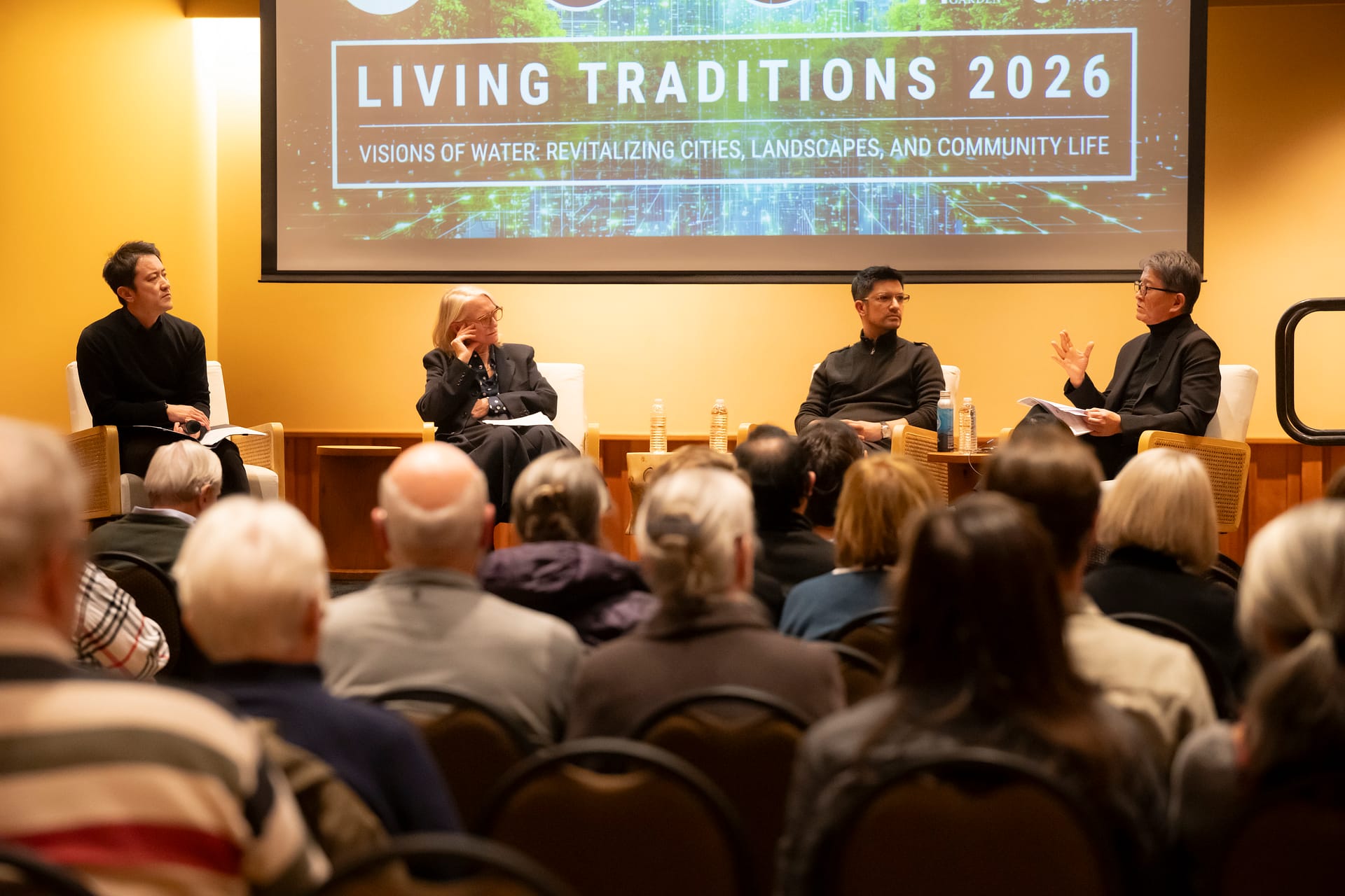 four people sitting down in chairs, chatting, in front of an audience