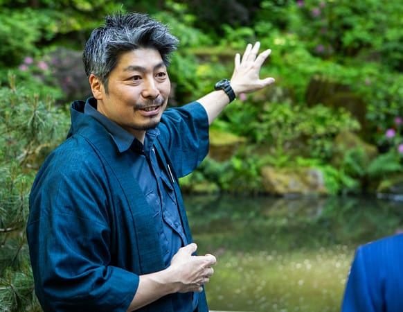 garden curator Hugo Torii stands smiling and gesturing to a waterfall wearing a Japanese gardening outfit and happi coat