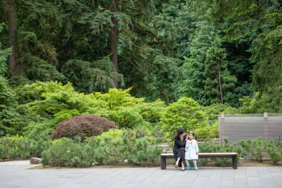 Adult woman and girl sitting on bench among the green foliage within the Japanese Garden.