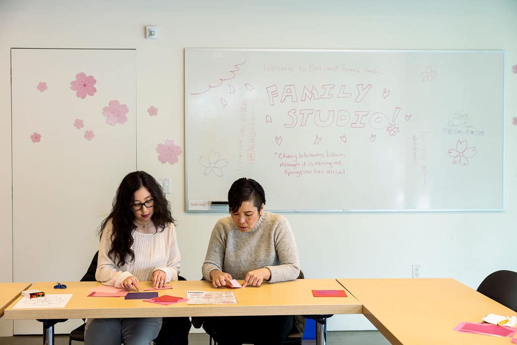 Two woman doing origami in front of a white board with the words "Family Studio"