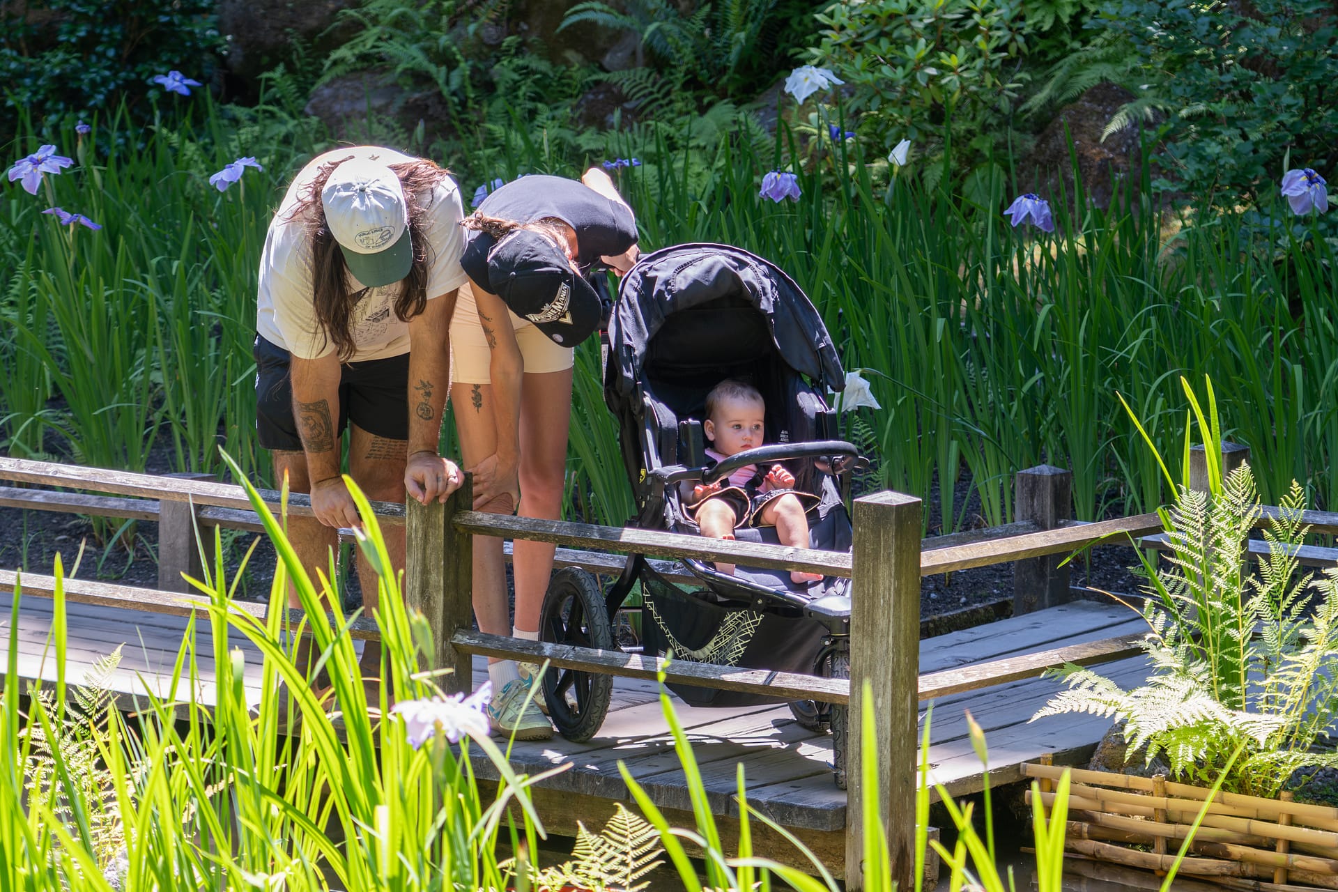 two parents lean over a baby stroller to look at their child on a wooden bridge