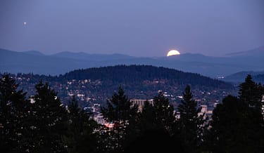 Bright moon rises over the hills behind a lit-city and forest foreground