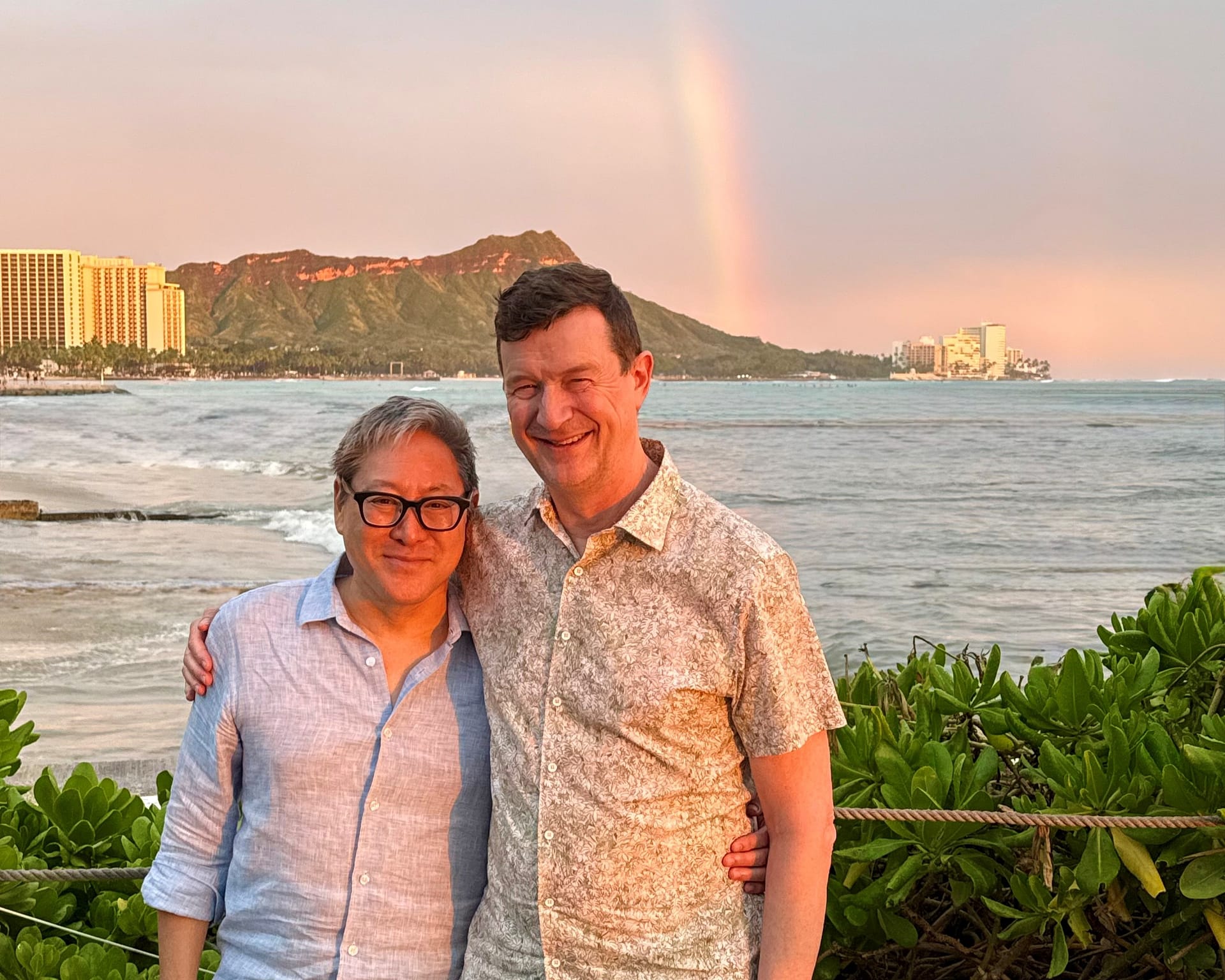 a couple stands in front of a body of water with a rainbow dipping down from the sky