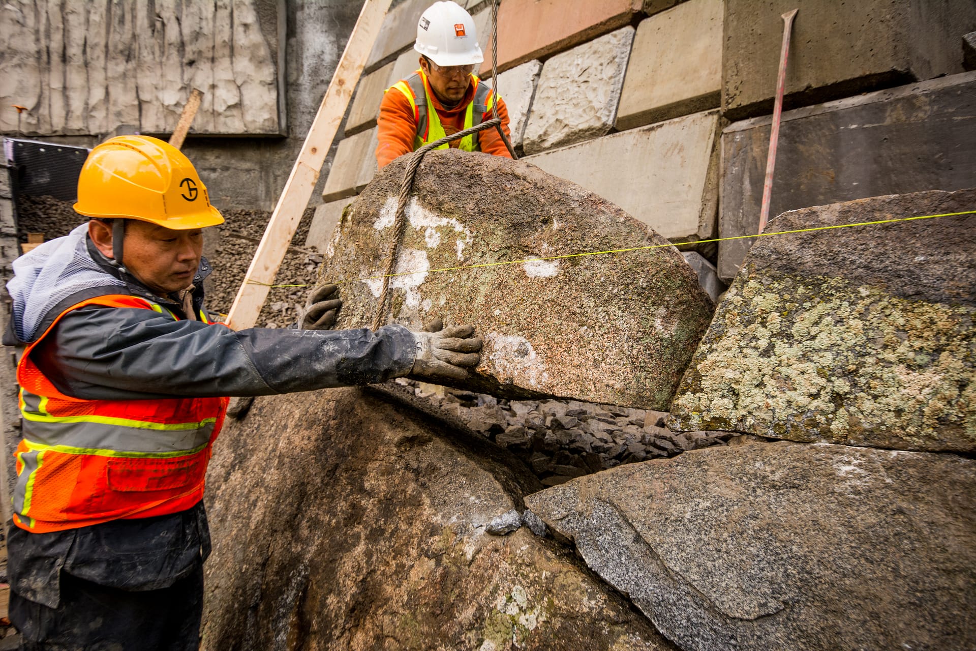 a person wearing a helmet and safety vest guiding a massive stone into place on a wall