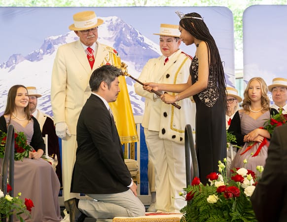A man kneels while being knighted by a young woman wearing a crown and holding a scepter.