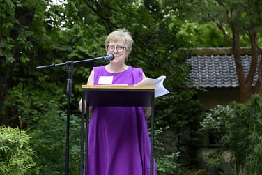 Caryl Stern stands at a podium in front of a wooden Japanese gate and trees.