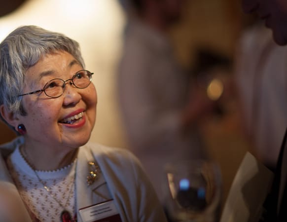 a woman with short gray hair and glasses smiles looking to someone off camera
