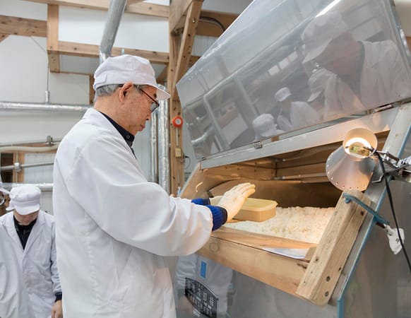 a man in a cot and hat holds a box over a stainless steel container of cooked rice