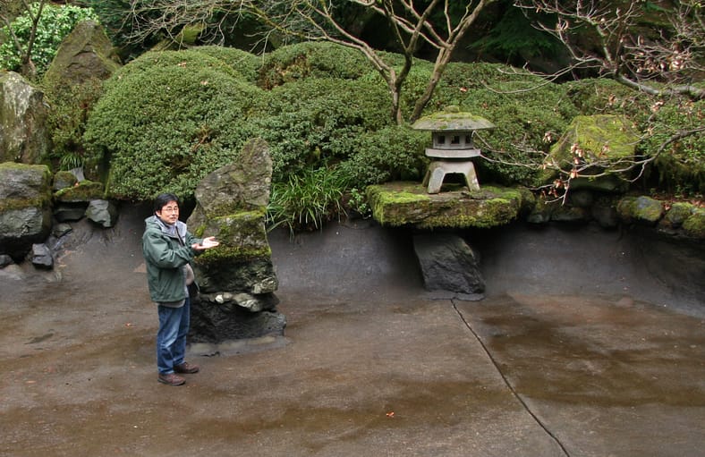 Sadafumi Uchiyama stands in a drained pond and gestures to a stone lantern.