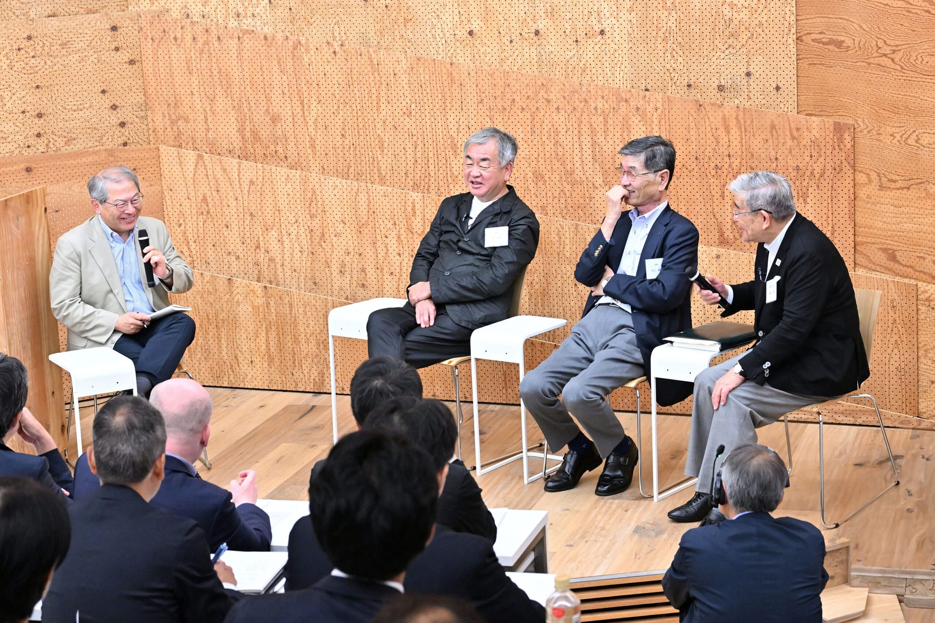 four men sit in front of a crowd in a wooden lecture hall having a moderated talk