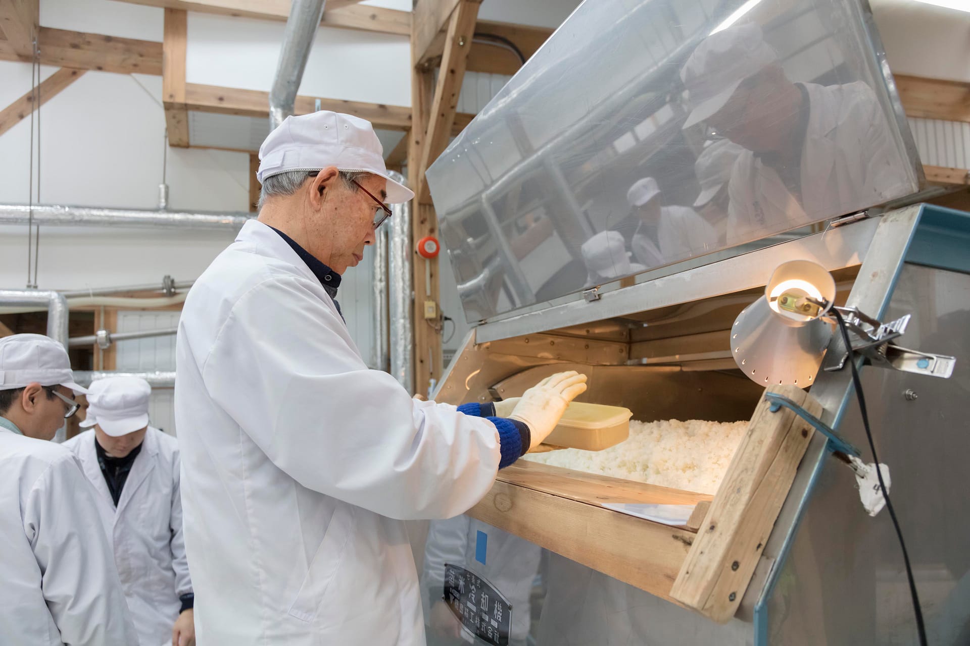 a man in a cot and hat holds a box over a stainless steel container of cooked rice