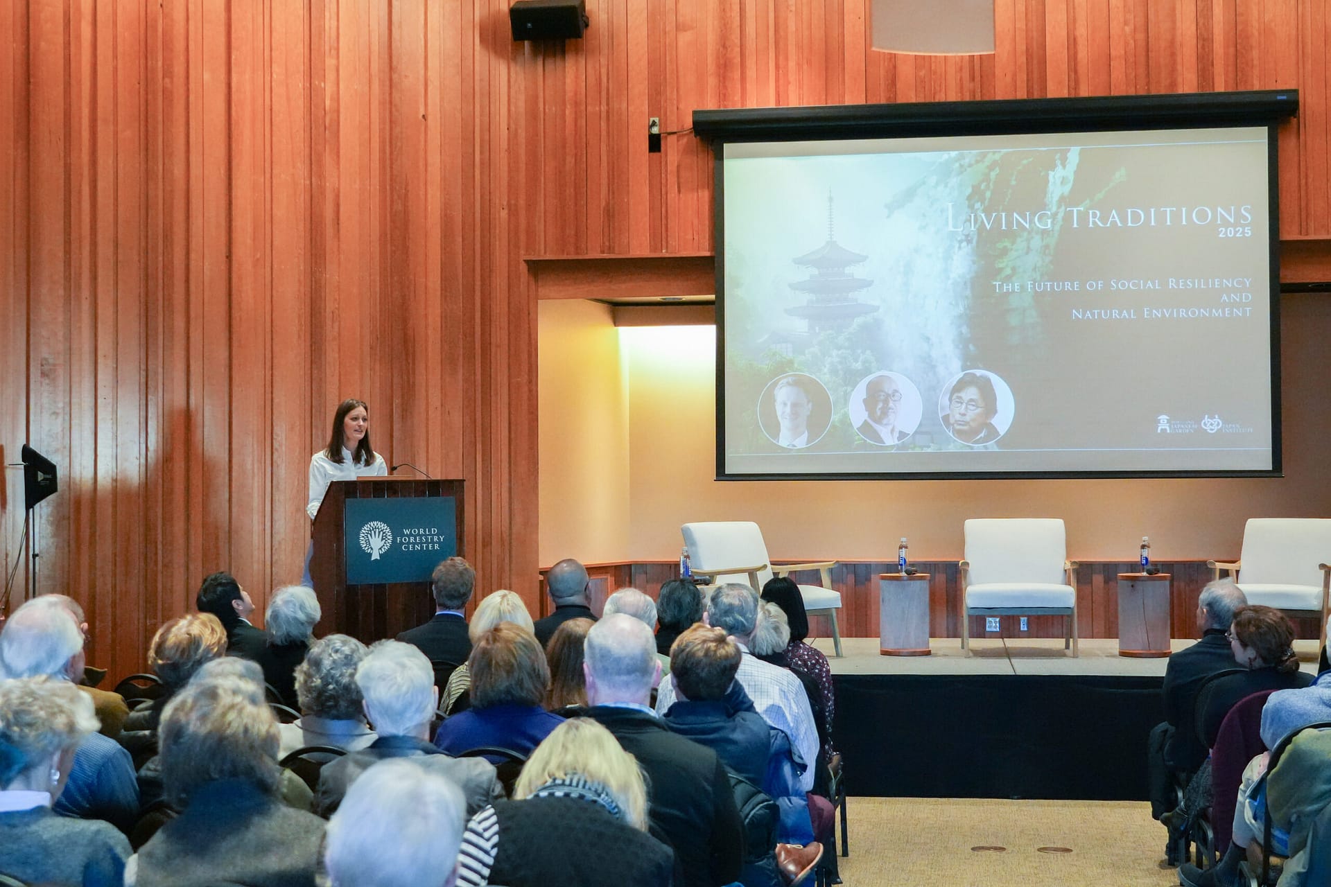 A woman stands behind a podium as an audience looks on.