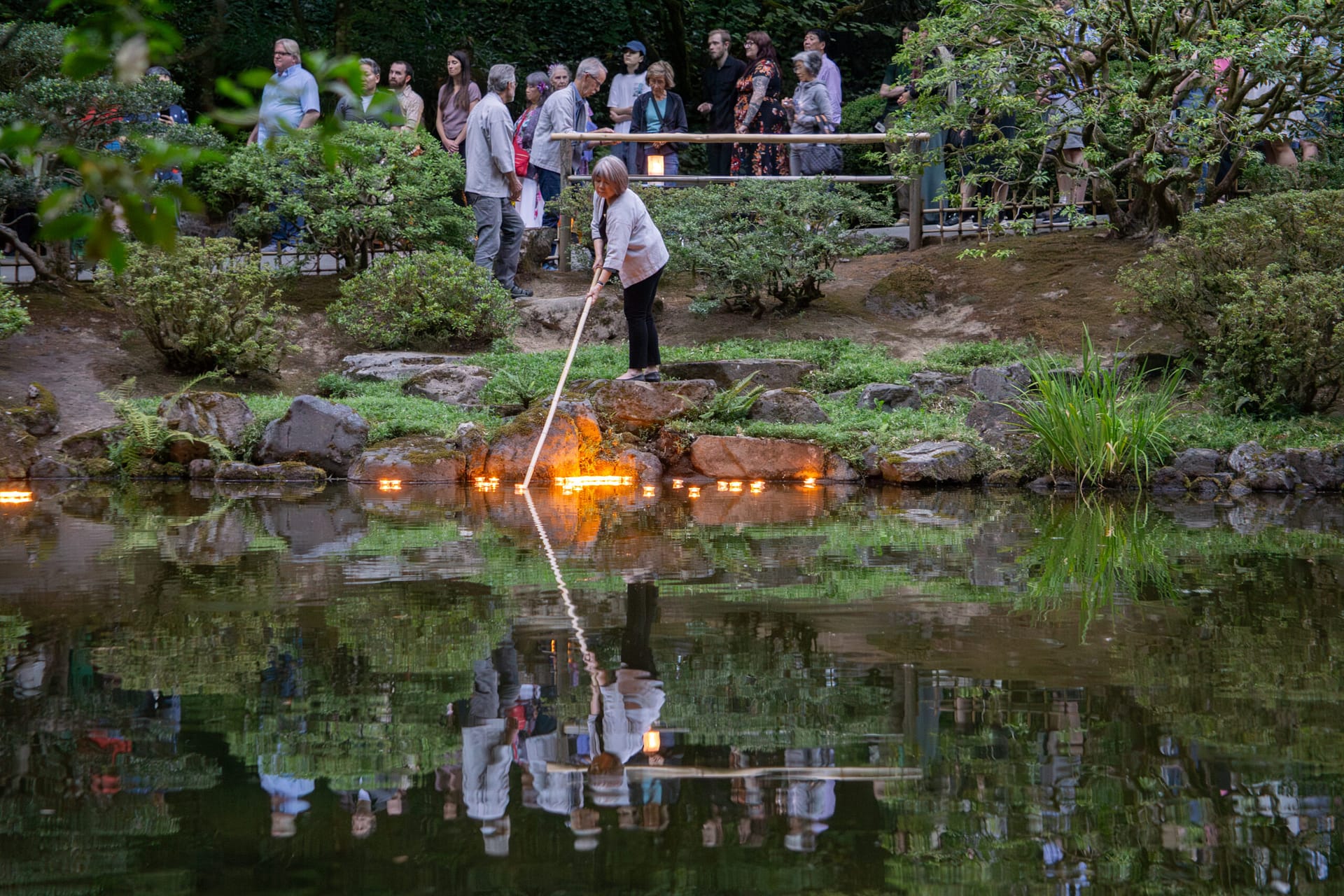 A woman pushes a candle into a pond using a long pole.