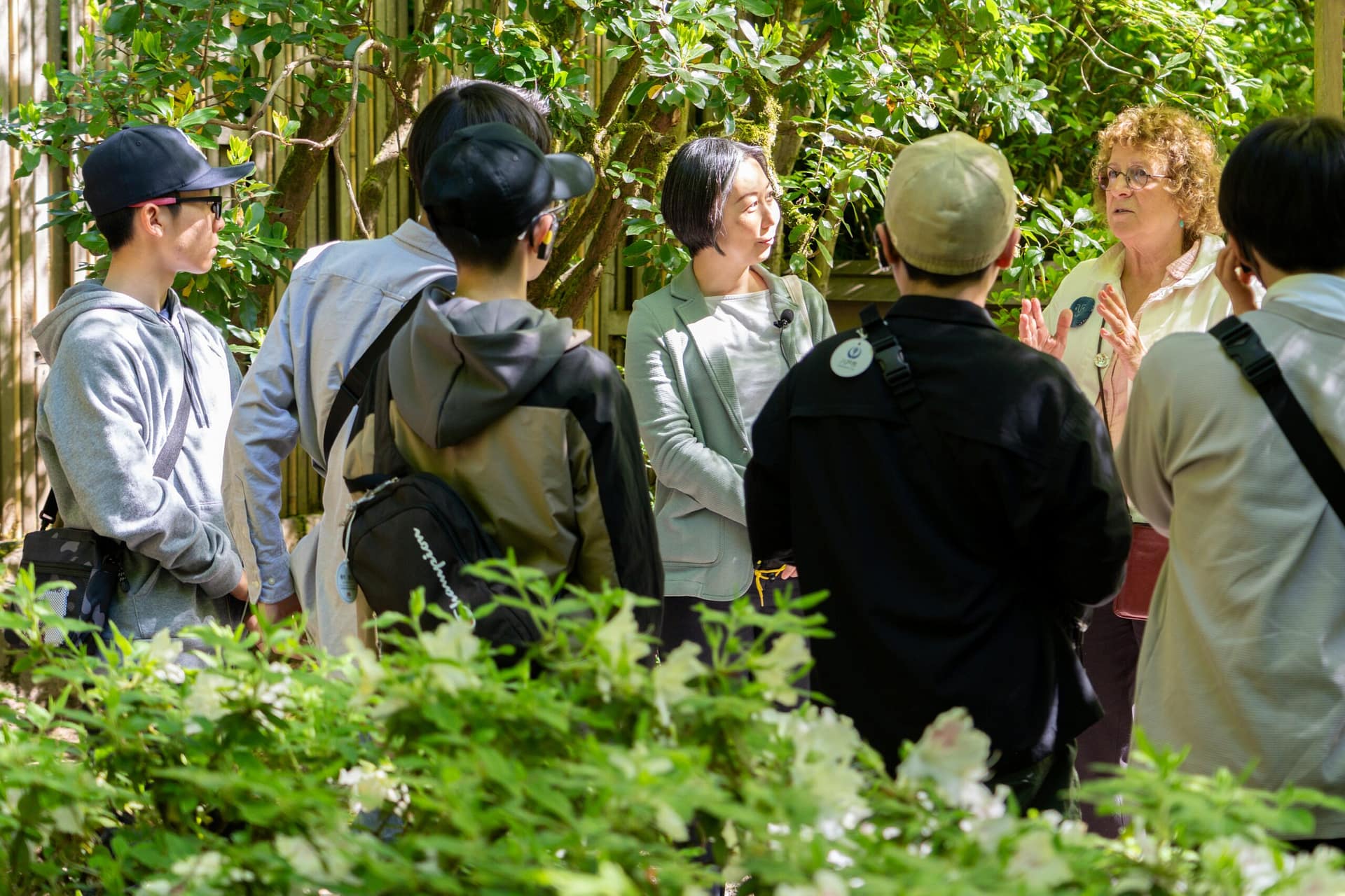 A group of young students listening to a woman in a garden.