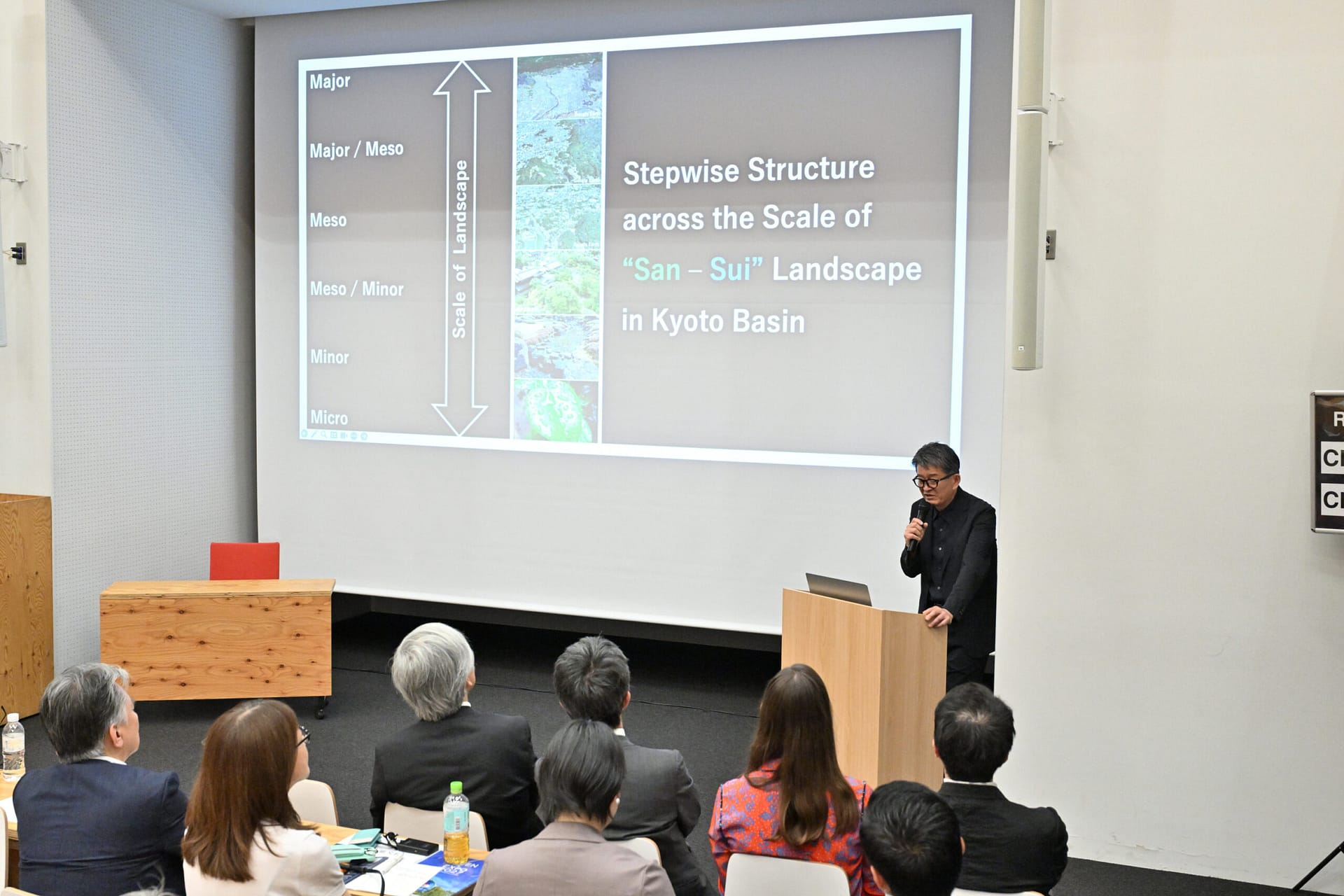 a man with a projection behind him that has different levels of scale: major, major/meso, meso, meso/minor, minor, and micro