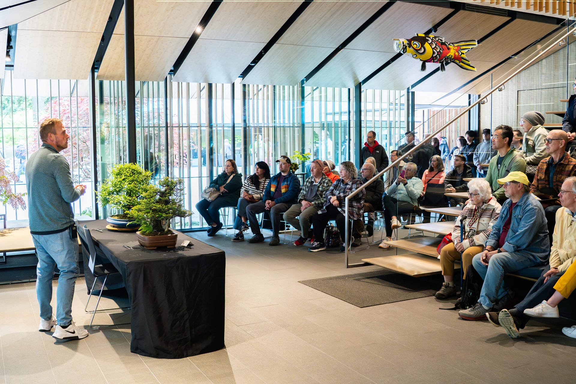 A seated audience watches a man talking about bonsai